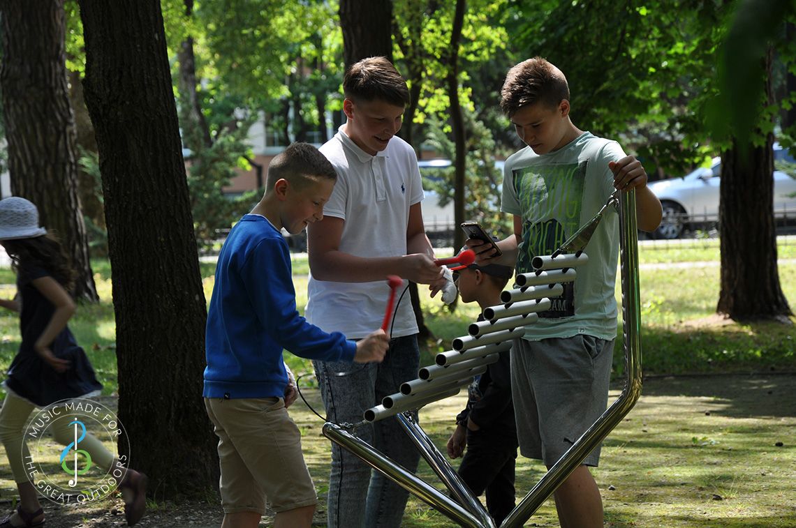 three teenage boys playing a large vertical xylophone made of metal in playground 