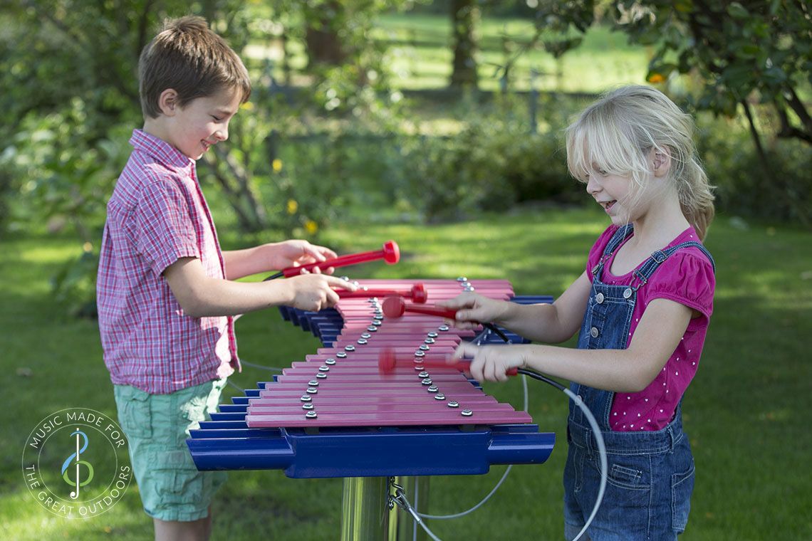 Boy and girl standing on opposite sides and playing a large blue outdoor xylophone with red beaters