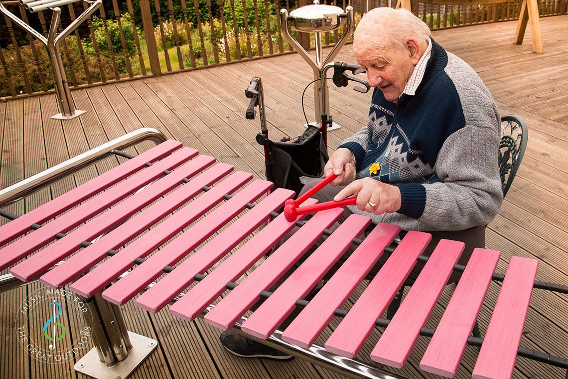 older man in wheelchair playing a large outdoor marimba in care home garden
