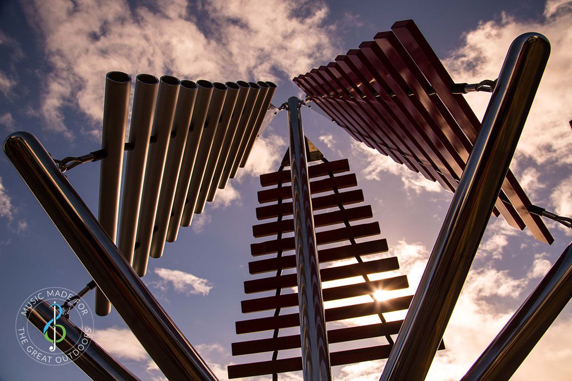 looking up from the ground at a large outdoor musical instrument with one stainless steel post in ground with three vertical xylophones attached