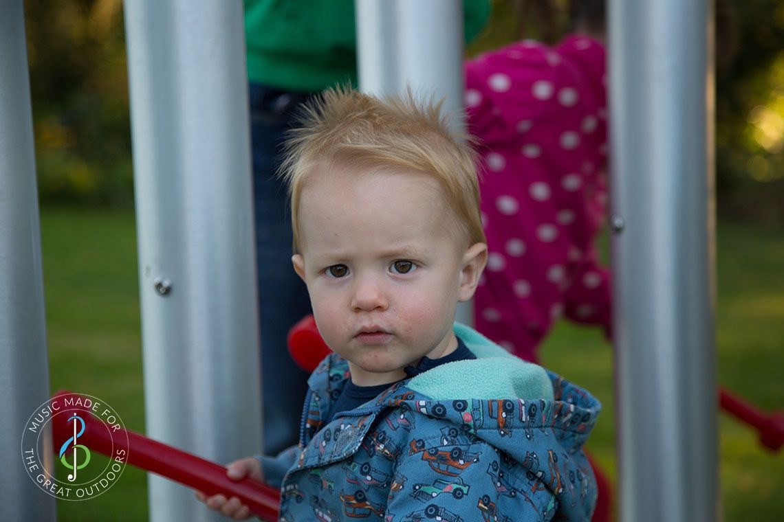 Close up of red haired baby boy hitting huge silver outdoor chimes with a red beater