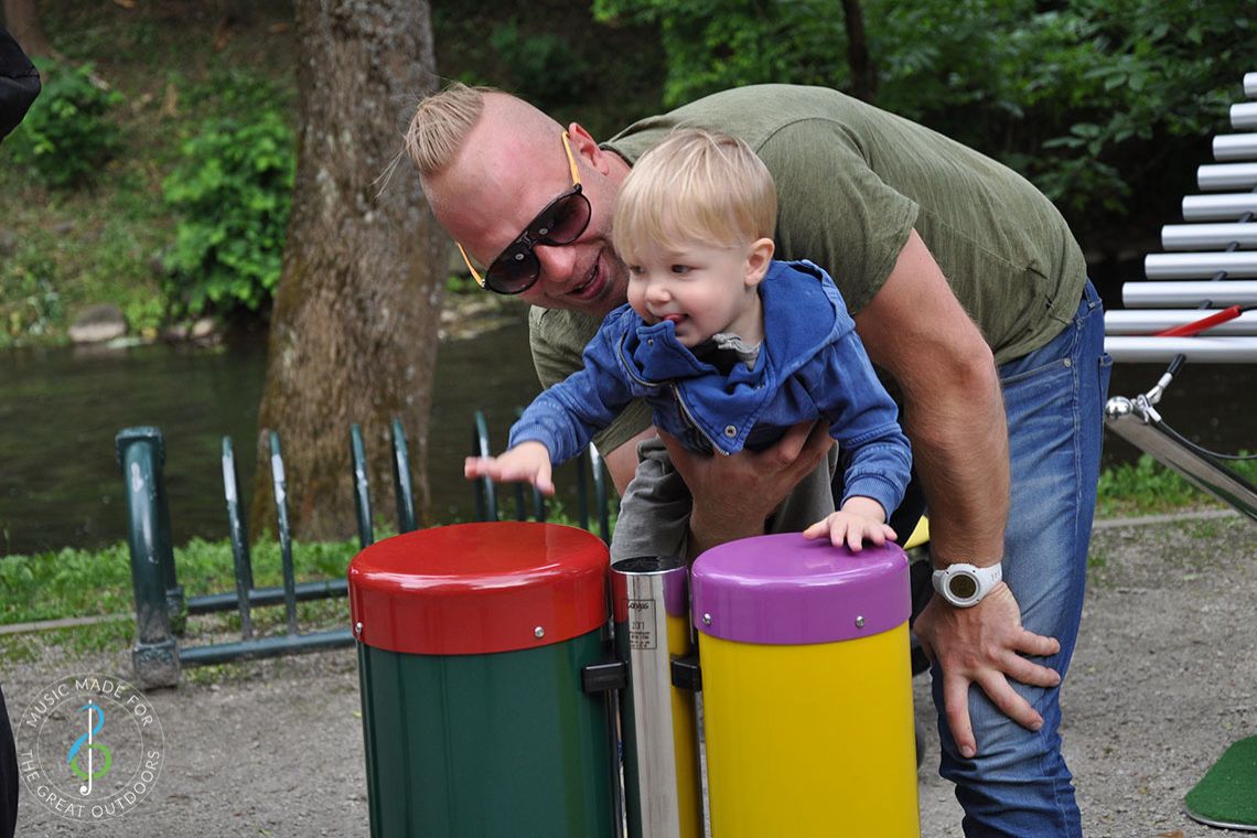 Toddler being held up by his father to play on colourful outdoor conga drums in park 